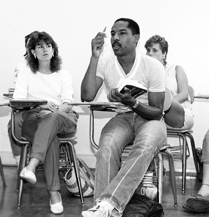 Image of three students sitting in class with a Black man in the front row raising his hand